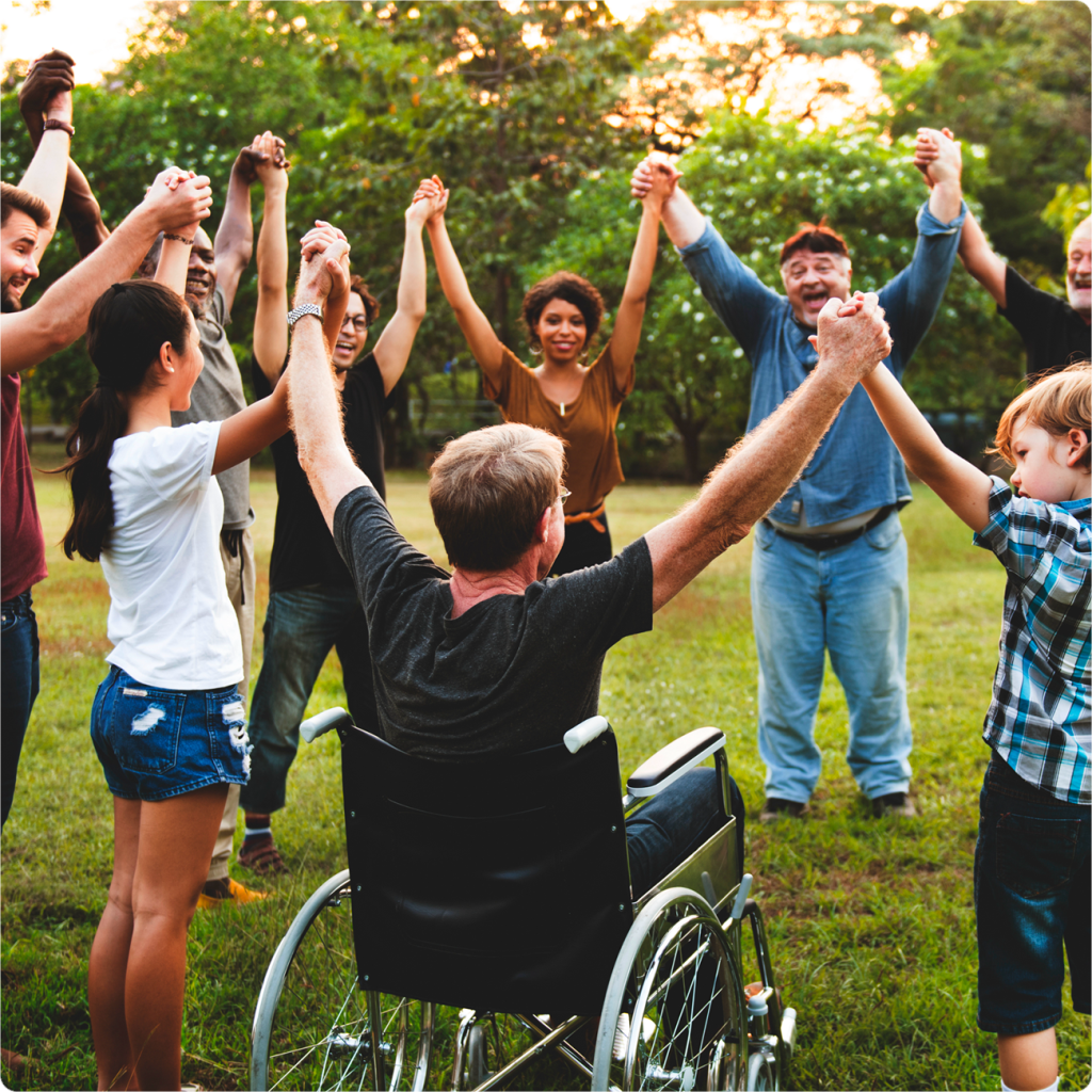 A diverse group of people holding hands in a circle, with a person in a wheelchair, outdoors in a park.