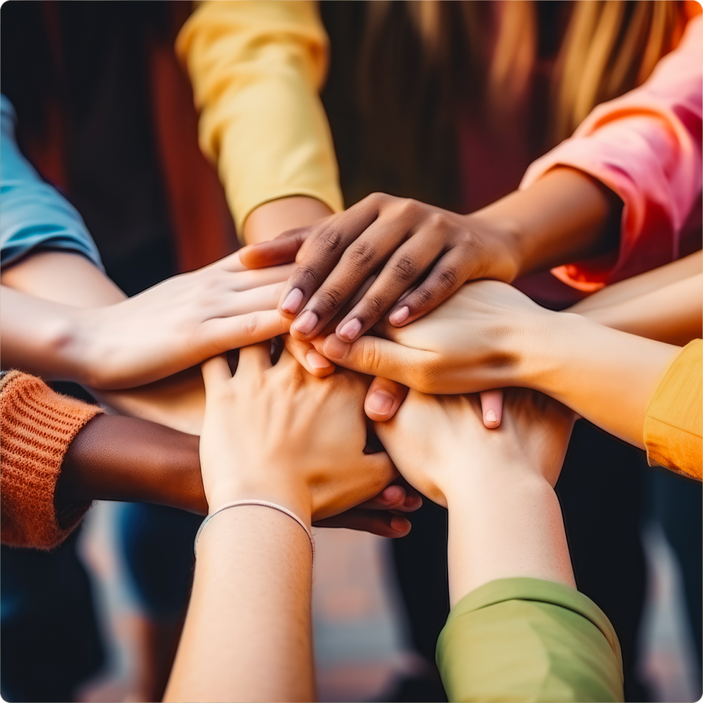 A group of people with diverse skin tones stack their hands together in a unified gesture; their colorful sleeves suggest individuality and teamwork.